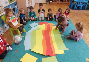 Dziewczynka pokazuje obrazek ze strefami klimatycznymi, a inna próbuje odwzorować go na dywanie.