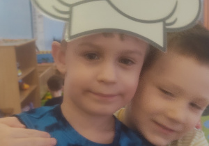 Dwóch chłopców przytula się do siebie, jeden z nich ma na głowie czapkę kucharza.